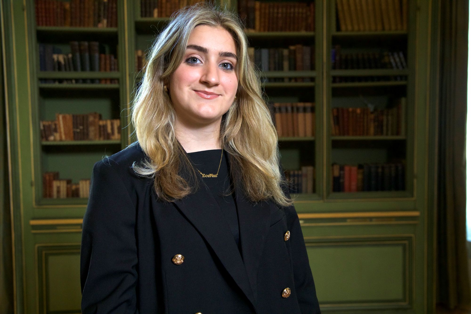 Person in a black blazer standing in front of a bookshelf filled with books, smiling at the camera.