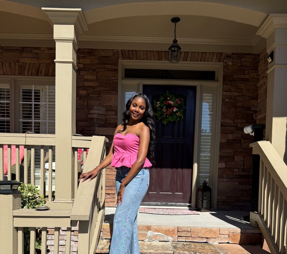 A person smiling while standing on a porch with a stone wall and a dark-colored door adorned with a wreath.