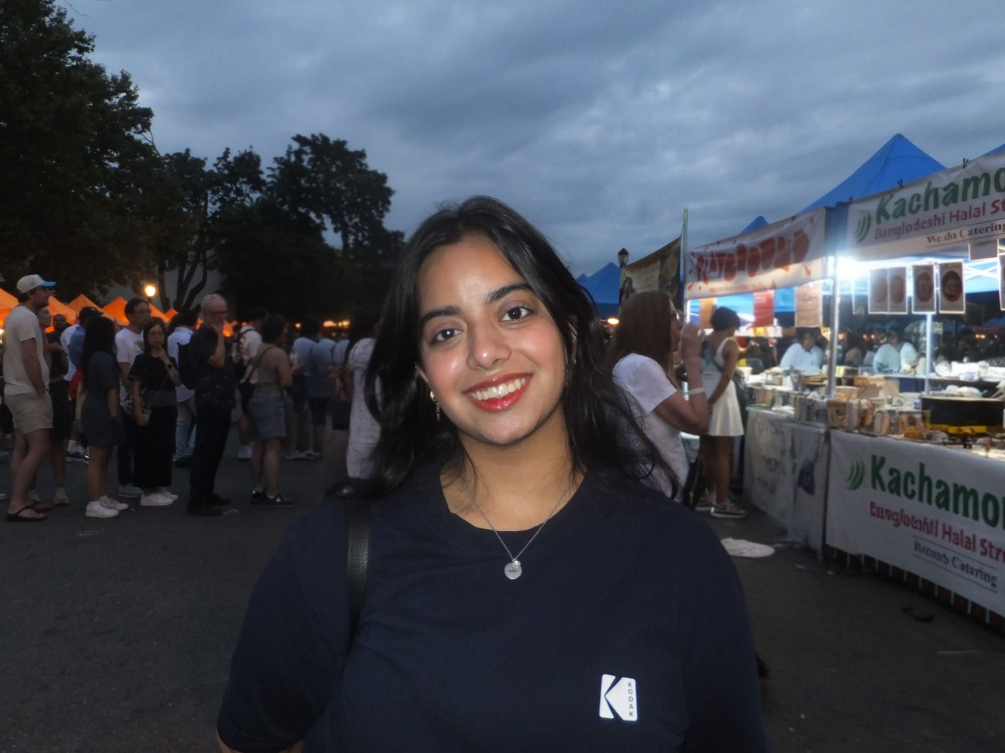 Individual smiling at a food festival with various stalls and people in the background during twilight.