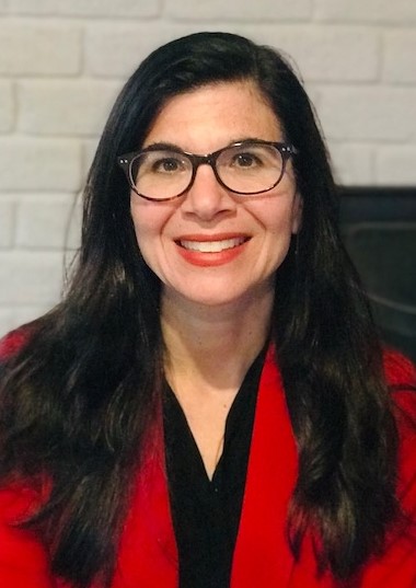 Female wearing glasses and red blazer poses in front of a white wall 