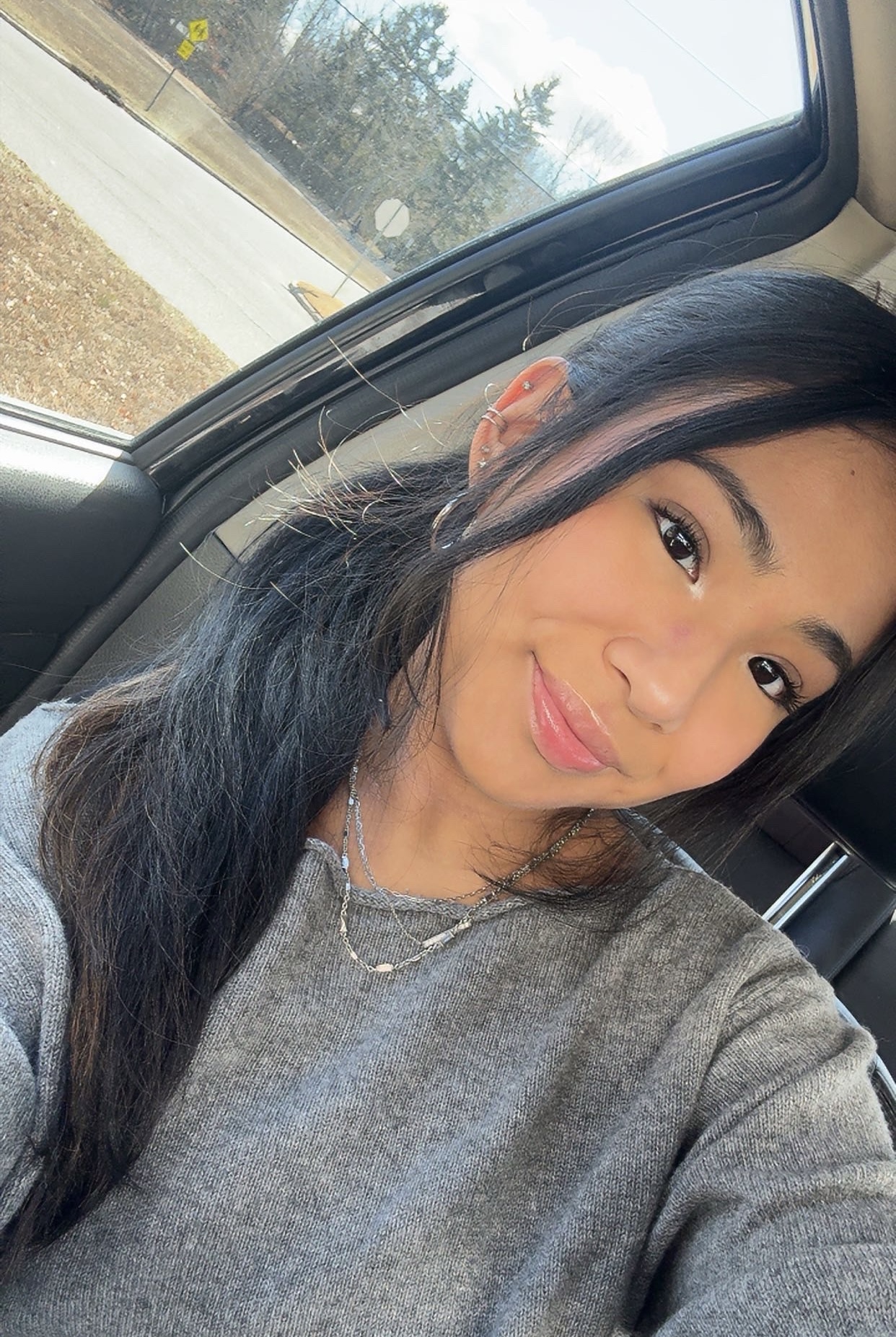 Person sitting in a car, smiling at the camera with sunlight filtering through the window, wearing a gray sweater and a necklace.