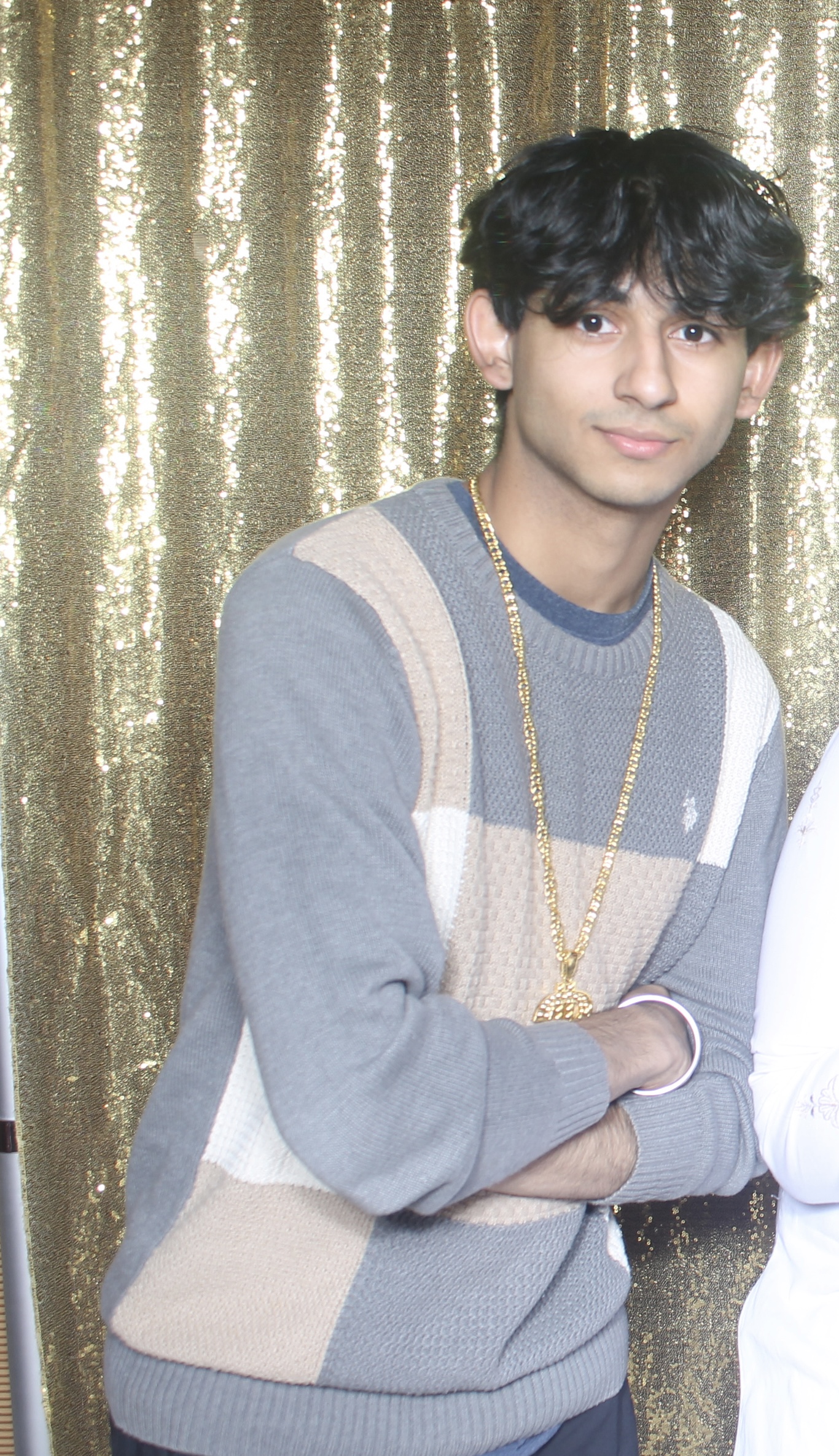 Person standing in front of a gold sequin backdrop, wearing a grey and beige striped sweater and a gold necklace.