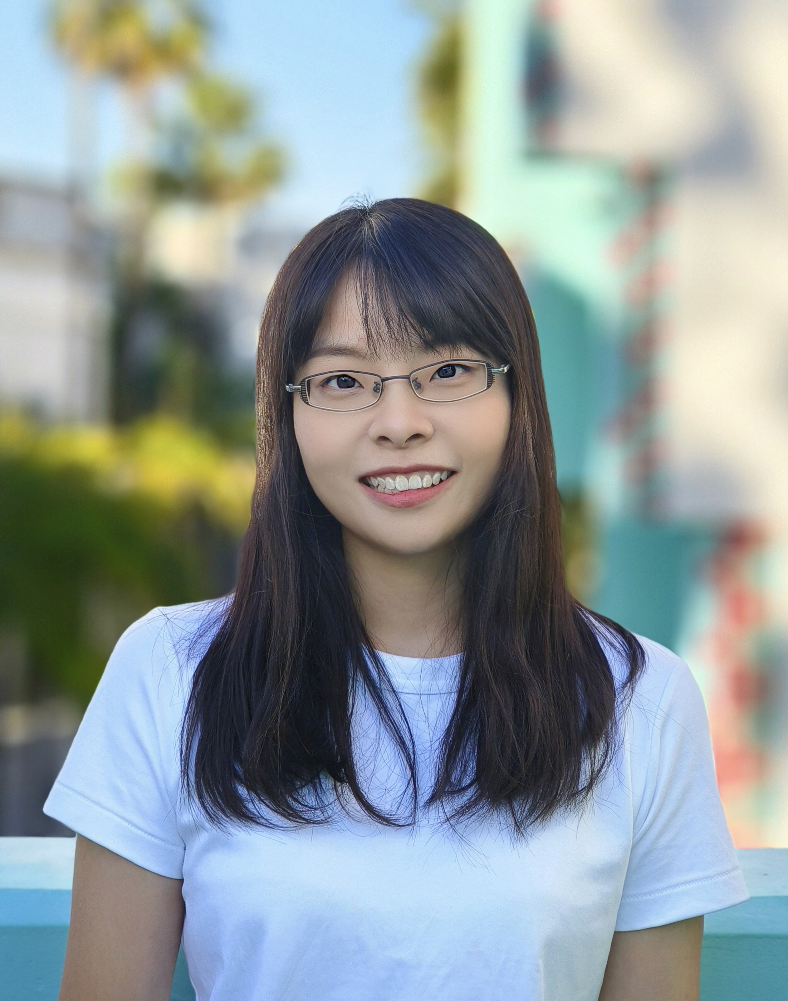 Portrait of a smiling person wearing glasses and a white t-shirt, standing outdoors with blurred trees and a blue sky in the background.