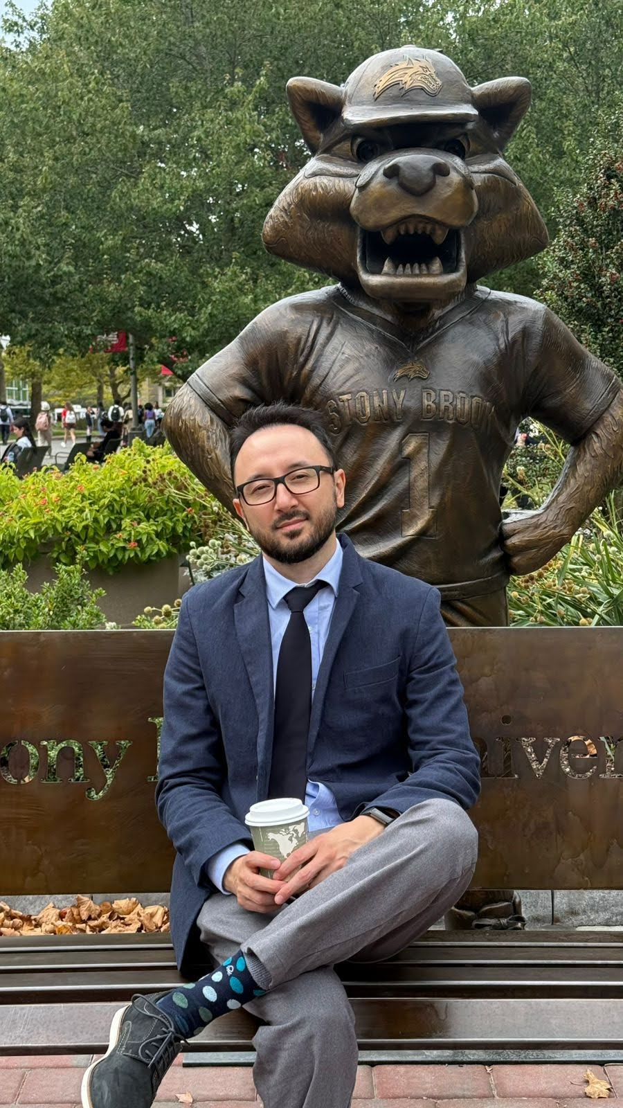 Person sitting in front of the Stony Brook Seawolf Statue, holding a coffee cup.