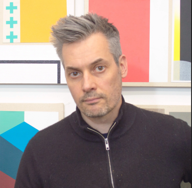 Person with gray hair and a black zip-up sweater standing in front of colorful abstract art.