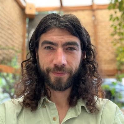 Portrait of a smiling person with long curly hair, wearing a light green shirt, outdoors.
