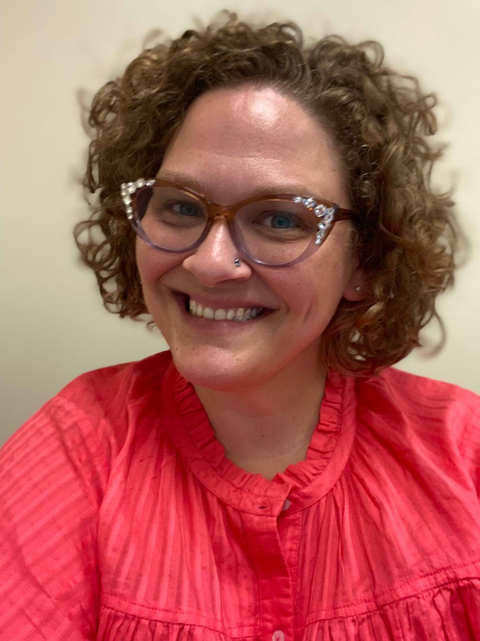 Person with curly hair and glasses smiling in a red shirt.