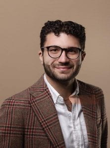 Portrait of a smiling person wearing glasses and a tweed jacket over a white shirt, set against a neutral background.