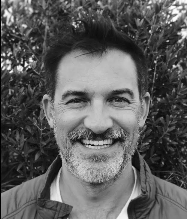 Close-up portrait of a smiling person with short hair and a beard, standing in front of a leafy backdrop.