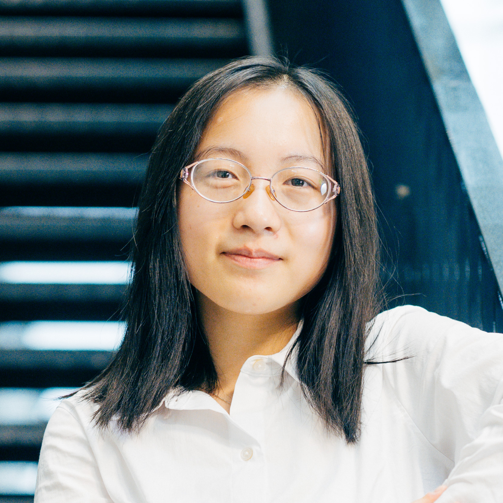A woman with glasses, wearing a white shirt, smiles subtly against a blurred dark stairway background, conveying confidence and calm.