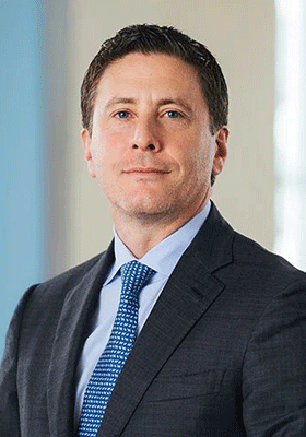 A confident man in a suit and blue tie stands against a blurred, light-colored background. He gazes into the distance, conveying professionalism.