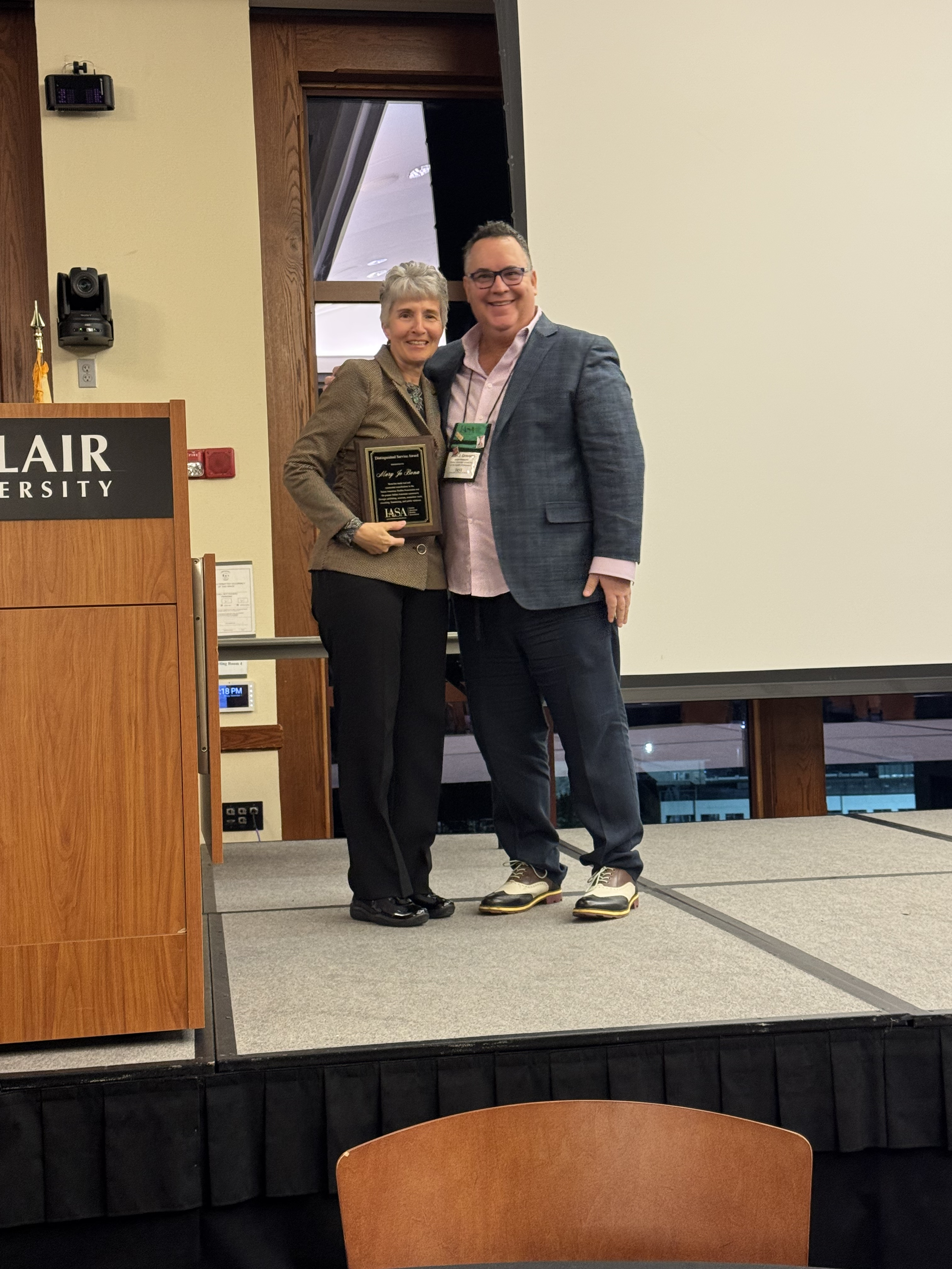 photo of Mary Jo Bona and Alan Gravano at the IASA conference. They're on a stage, by a podium, and Bona is holding her award