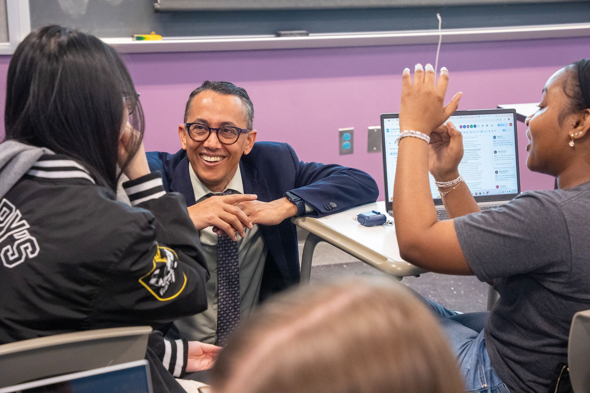A smiling faculty member in a suit and tie kneels between two students who are sitting at their desks in a classroom.