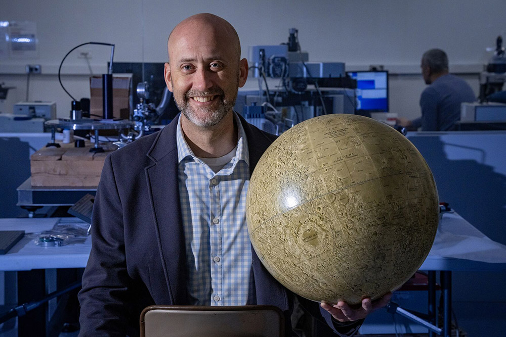 Timothy Glotch Timothy Glotch wearing a blue and white checkered shirt and blue blazer holds a moon model standing in a lab