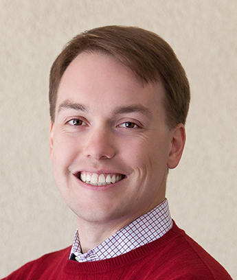 Portrait of a smiling person wearing a red sweater over a checked shirt, set against a neutral-colored background.