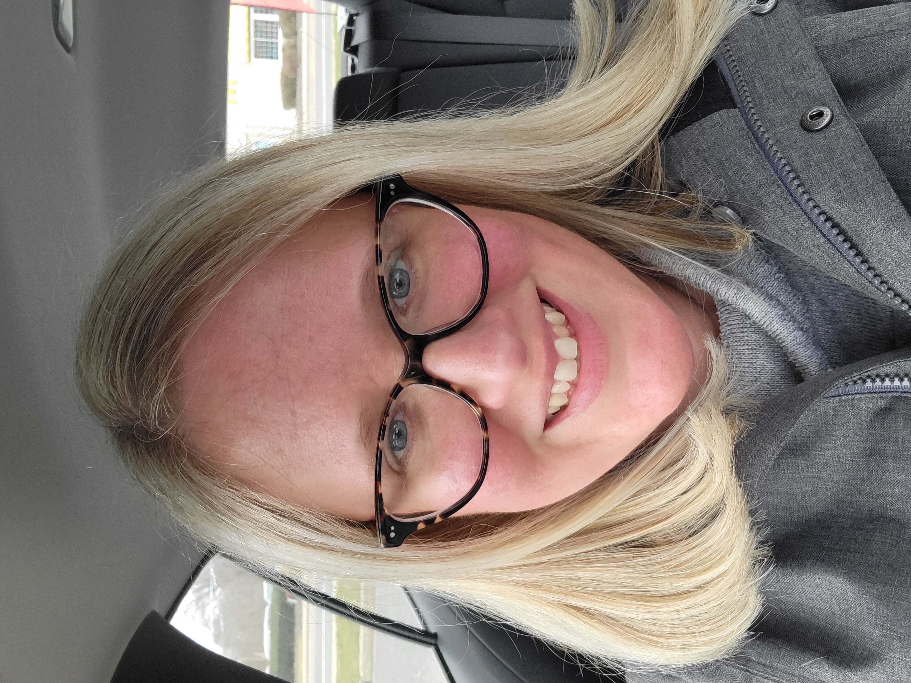 Close-up photo of a smiling person with blonde hair wearing glasses, inside a car.