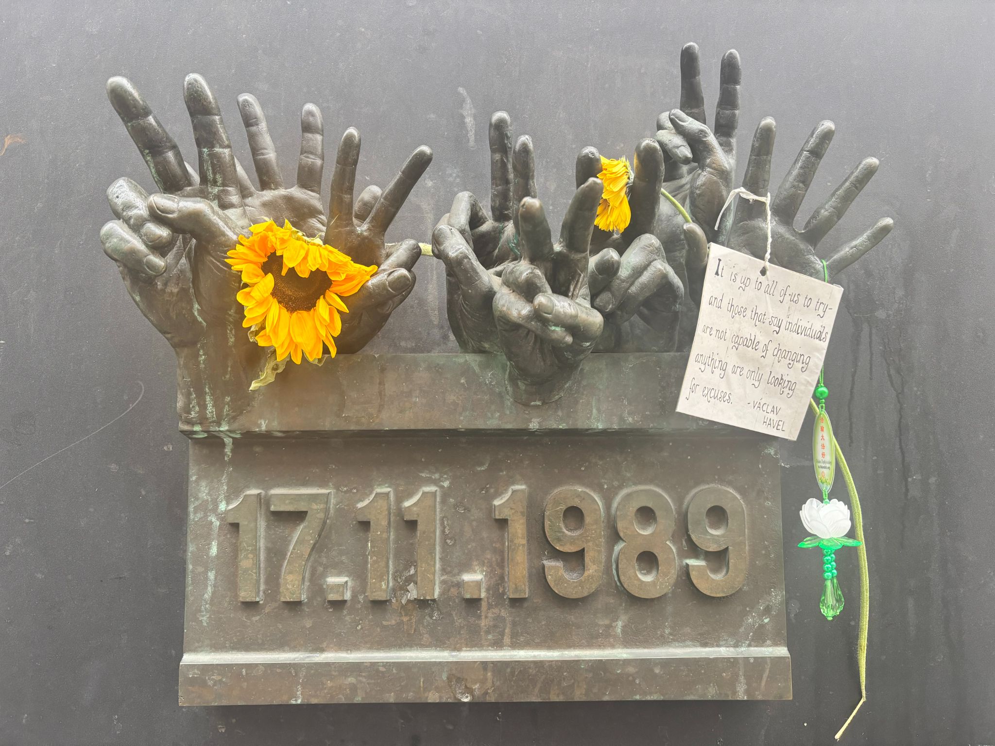 Bronze sculpture of multiple hands reaching upwards, each adorned with yellow flowers and a green four-leaf clover. A plaque with the dates "17.11-1989" and a note with a message are also visible.