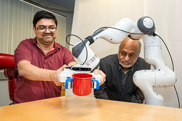 Two men interact with a robotic arm gripping a red mug on a table. One man smiles while the other observes, showcasing technology and human interaction.