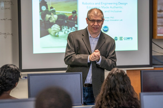 A man in a suit is giving a lecture in a classroom, standing in front of a projector displaying a slide about robotics and engineering design. Students are seated at computers, listening attentively.