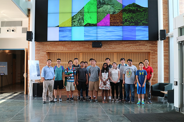 Computer Science and Informatics Summer Research Experience Program A group of 16 people stands in a line indoors, smiling. Behind them, a large, colorful digital mural depicting a mountain and water is displayed. The setting is bright and modern.