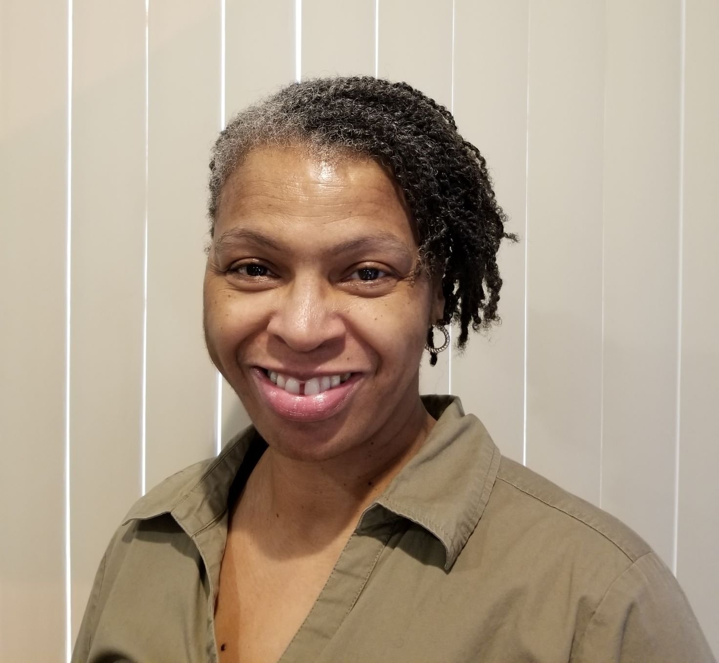 A headshot of a smiling woman with salt-and-pepper hair styled in short twists. She is wearing small hoop earrings and an olive-green button-down shirt. The background consists of light-colored vertical blinds.