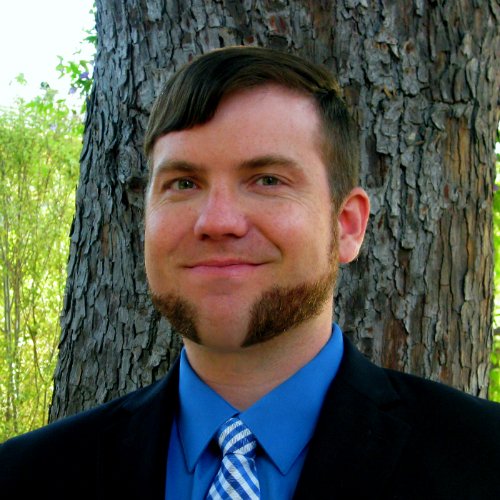 A portrait of a man with short brown hair and a distinctive mutton chop beard. He is smiling and dressed in a black suit jacket, a bright blue dress shirt, and a blue and white striped tie. He is standing outdoors in front of a large tree with textured bark.