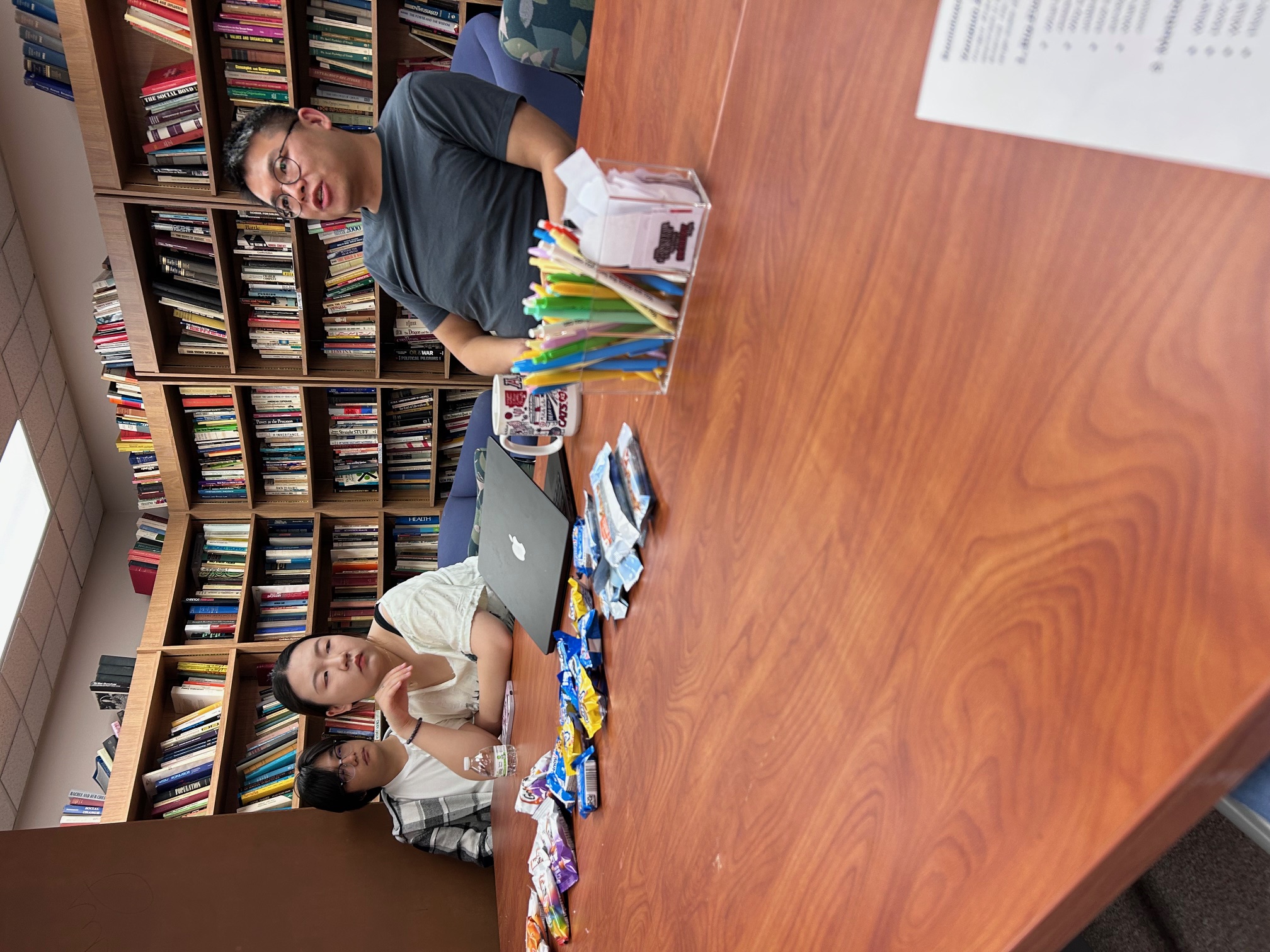 Three people seated at a conference table with snacks, a laptop, and bookshelves in the background.